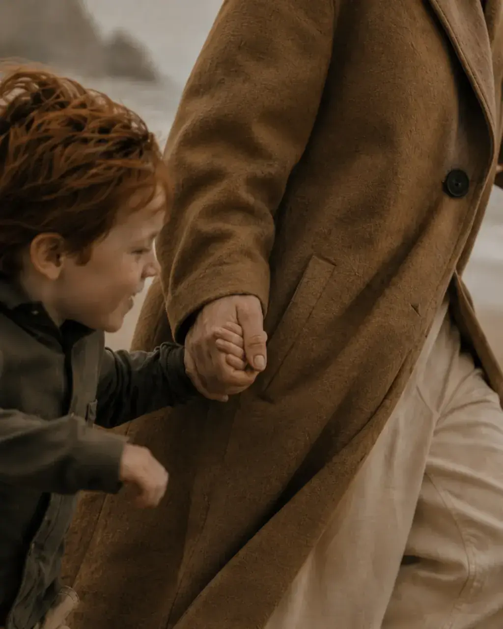Enfant roux tenant la main d'un adulte en manteau caramel – gros plan mains sur plage