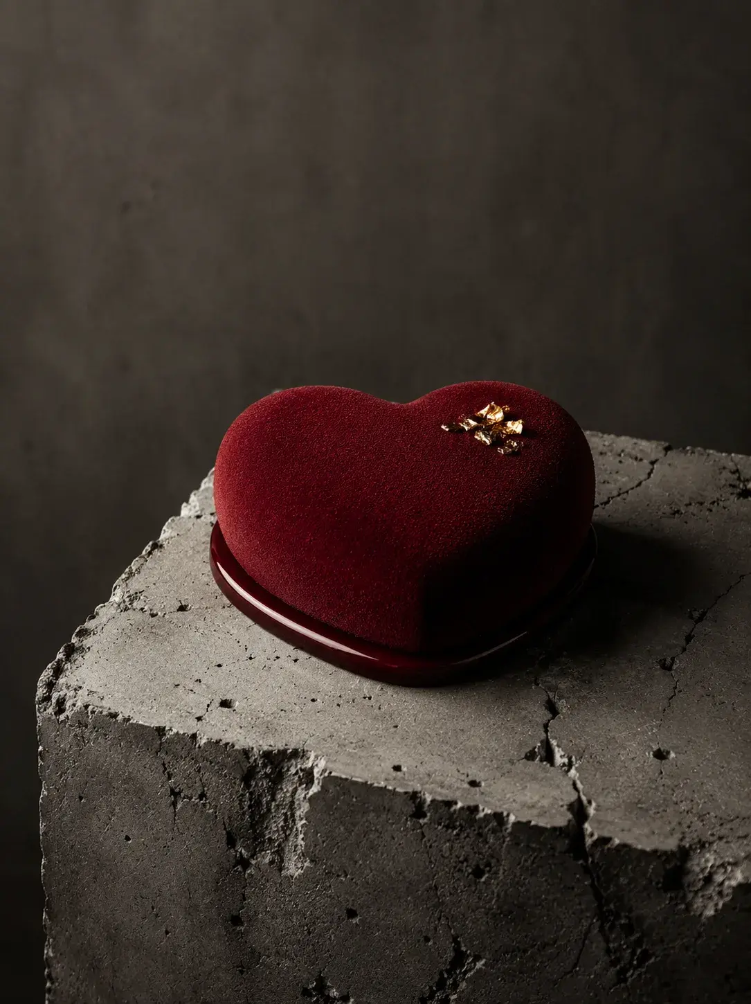 Entremets cœur velours rouge bordeaux sur bloc béton – éclats feuille d'or, fond gris sombre