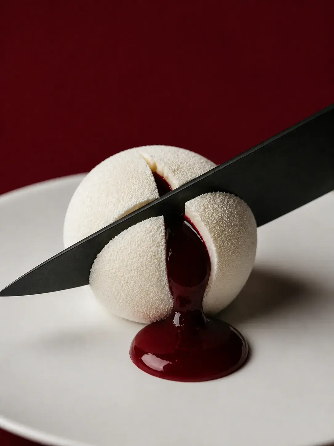 Entremets sphérique velours blanc sur assiette – couteau noir traversant, coulis rouge coulant