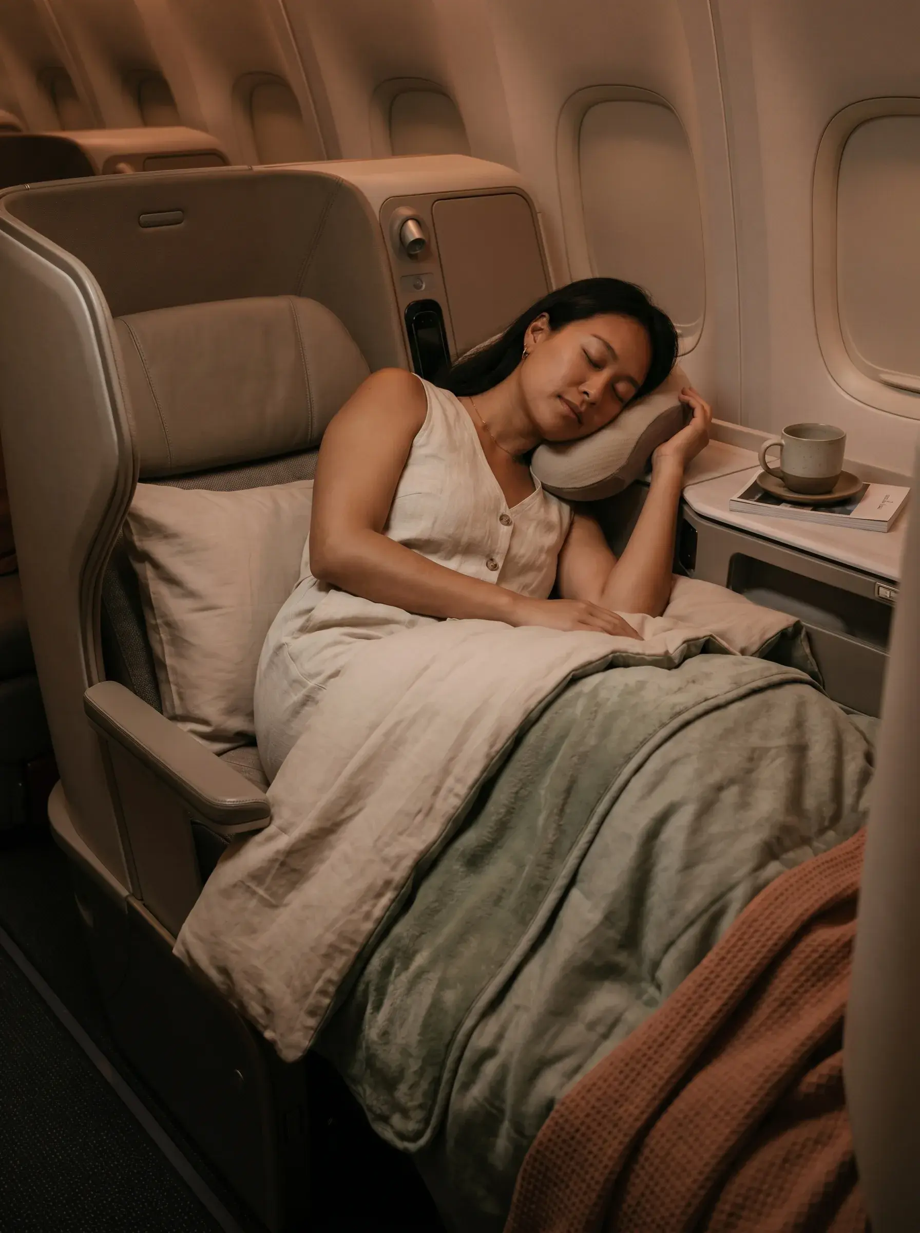 Femme asiatique dormant en classe affaires d'avion, siège totalement allongé, couette et coussin vert, ambiance feutrée