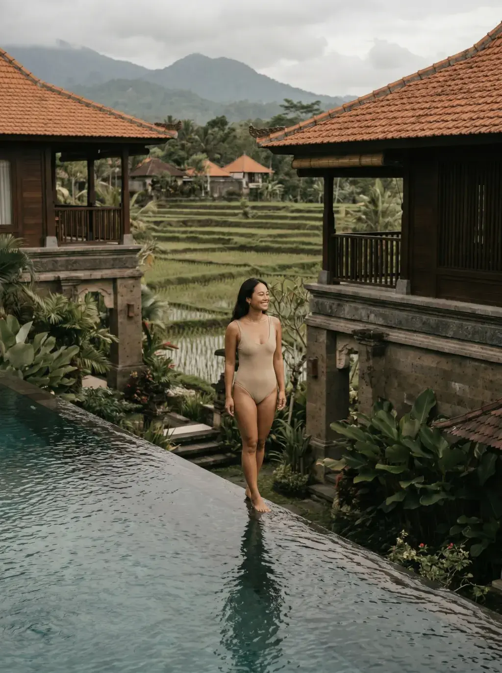 Femme asiatique en maillot de bain beige marchant au bord d'une piscine à débordement de villa balinaise, rizières en terrasses et montagnes en fond