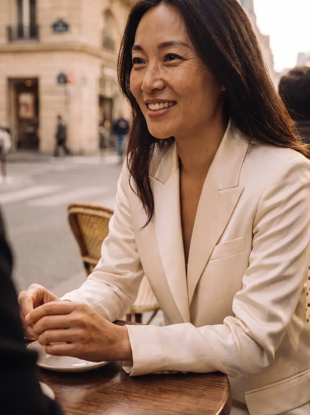 Femme asiatique souriante en blazer blanc en terrasse de café parisien