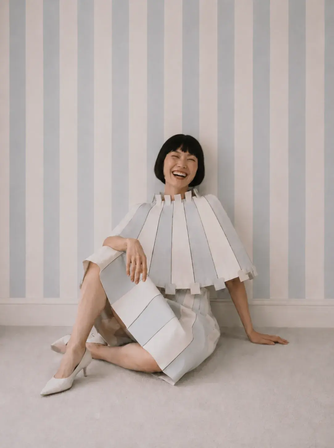 Femme assise au sol en cape origami riant – papier peint rayé bleu et blanc