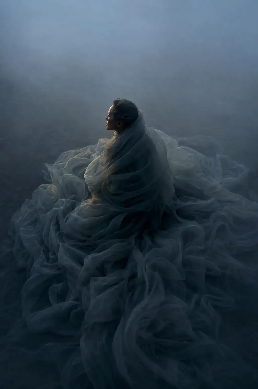 Femme assise enveloppée dans voile gaze bleu-gris – fond brumeux, posture méditative