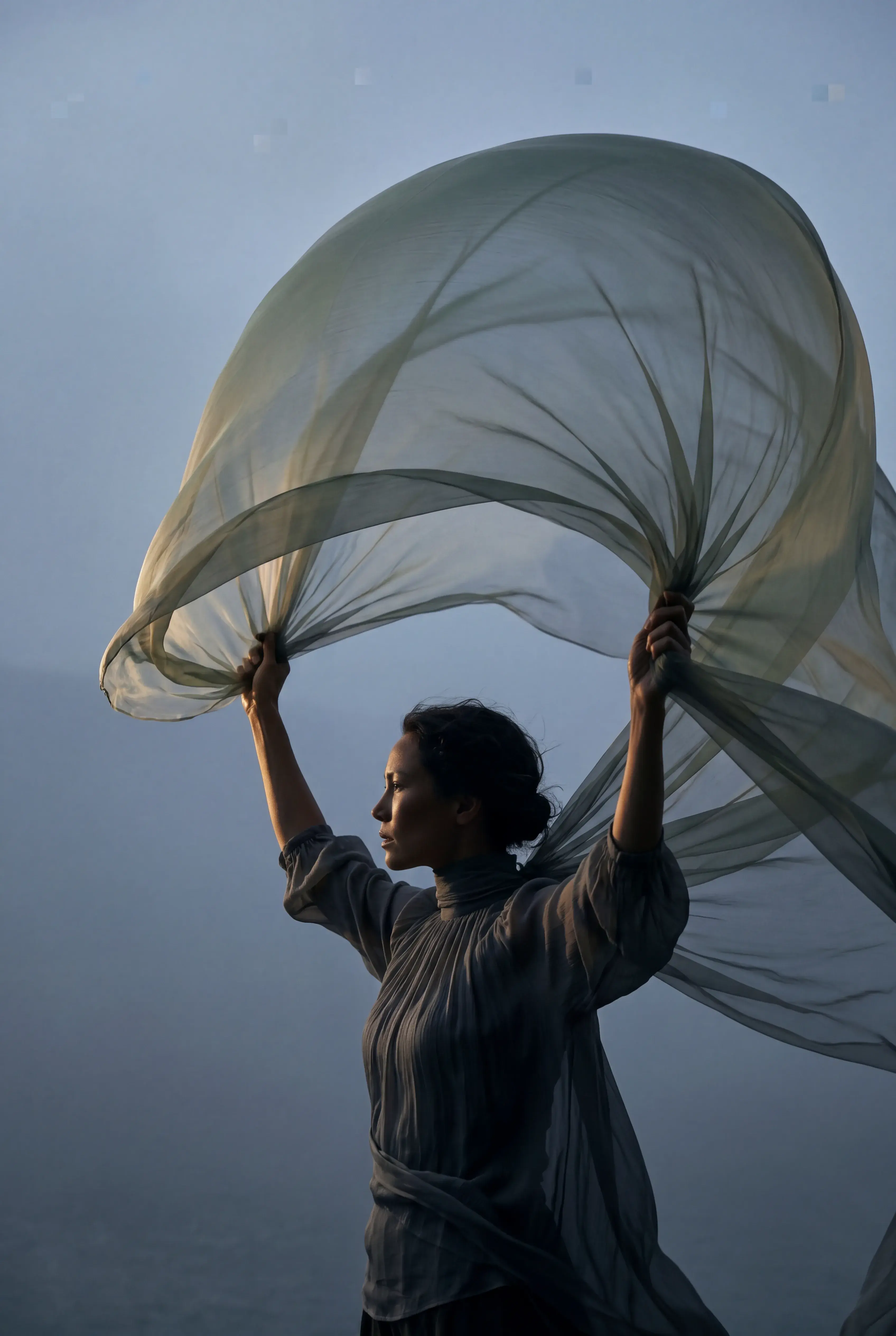 Femme bras levés tenant grand voile gaze bleu-vert – brume, ciel gris, posture de liberté