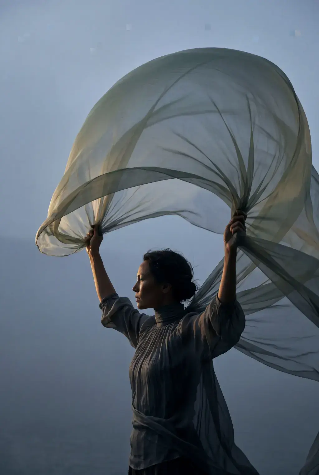 Femme bras levés tenant grand voile gaze bleu-vert – brume, ciel gris, posture de liberté