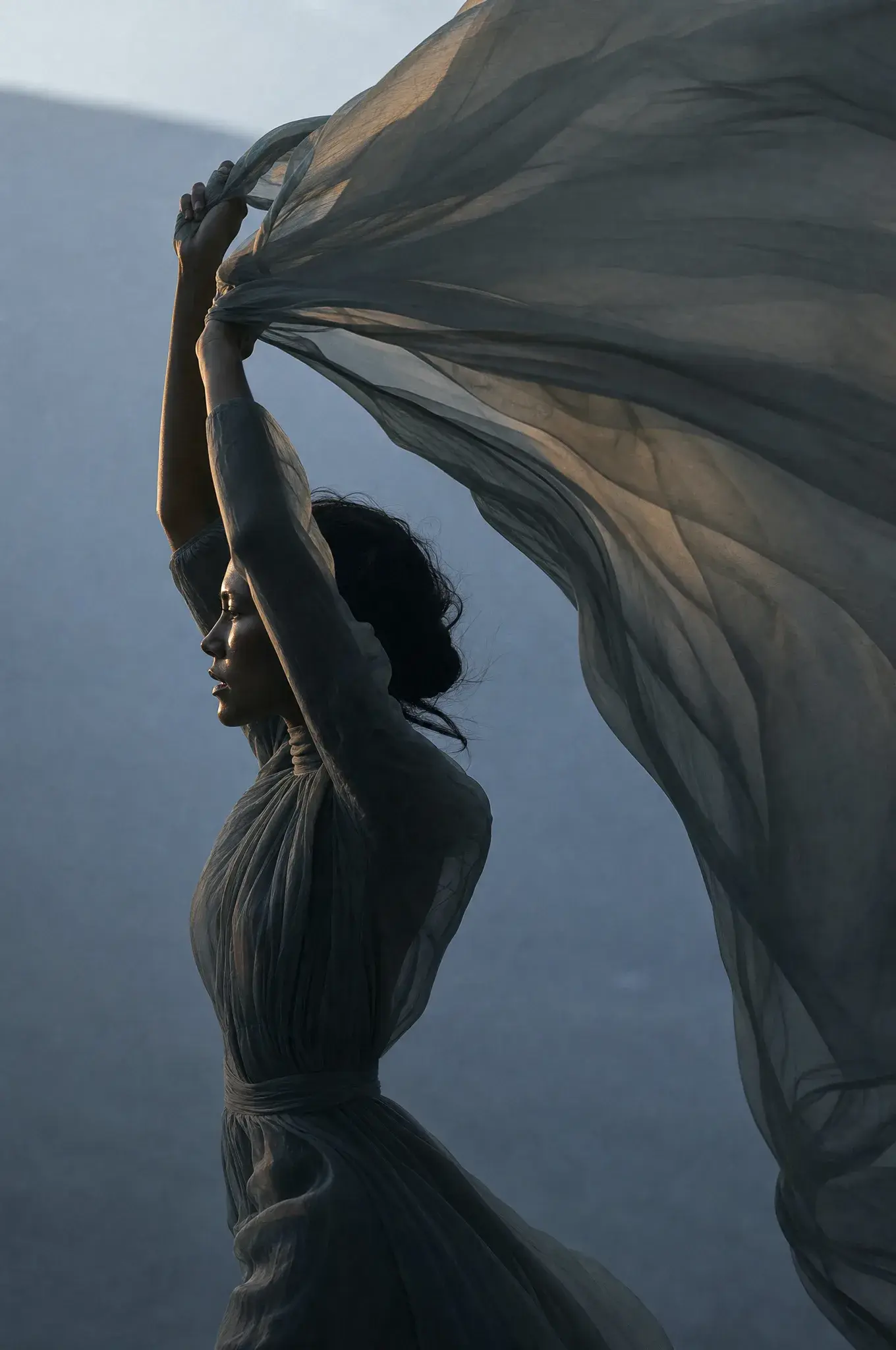 Femme bras levés tenant voile gaze flottant – silhouette contre montagne brumeuse, lumière dorée
