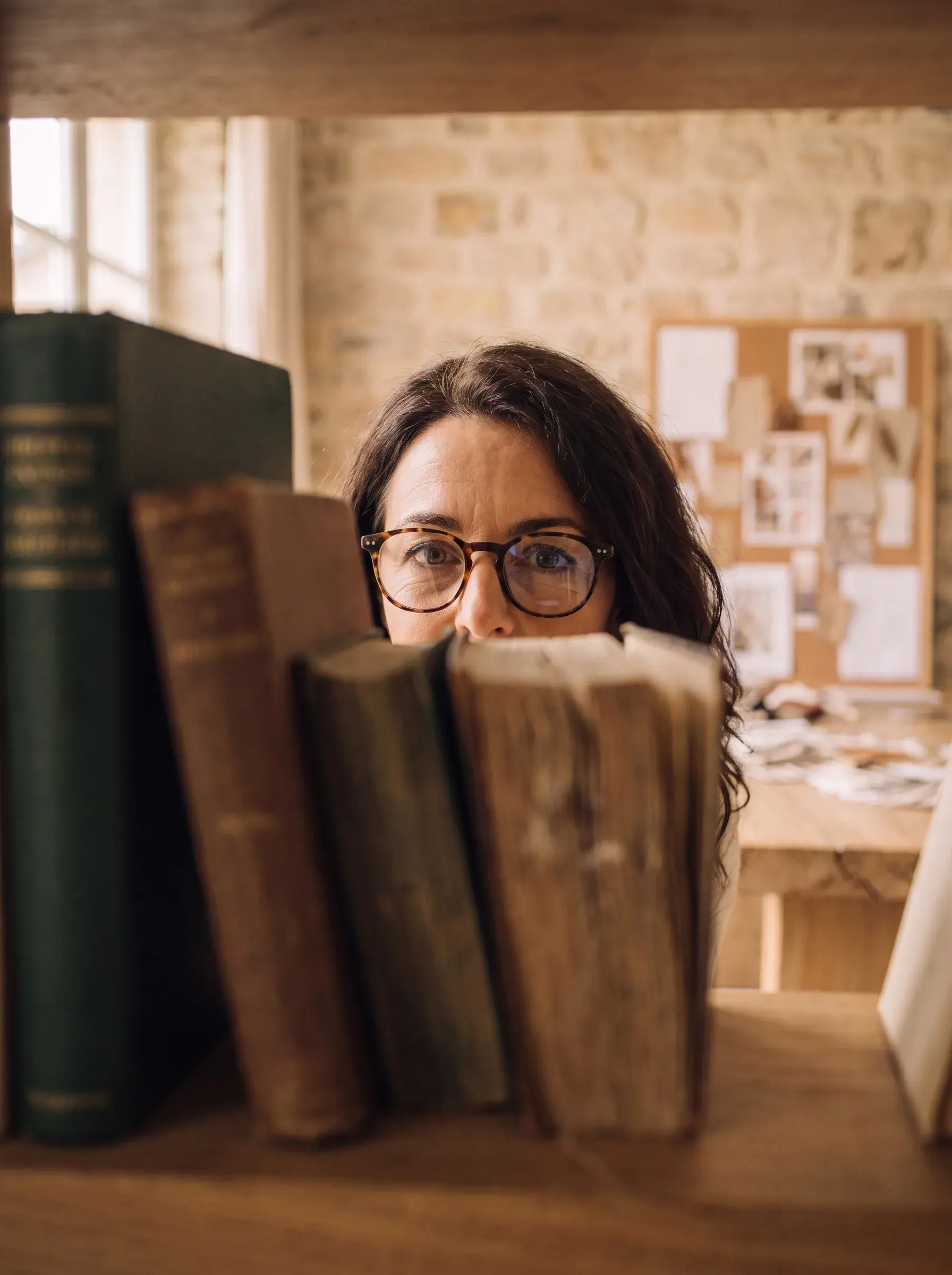 Femme brune à lunettes rondes épiant à travers des livres anciens sur une étagère