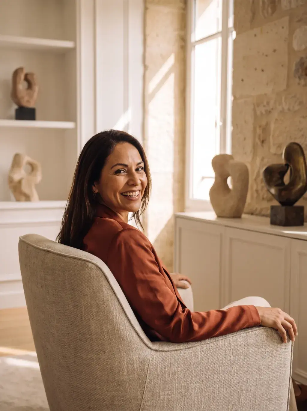 Femme brune en blazer terracotta assise dans un fauteuil avec des sculptures