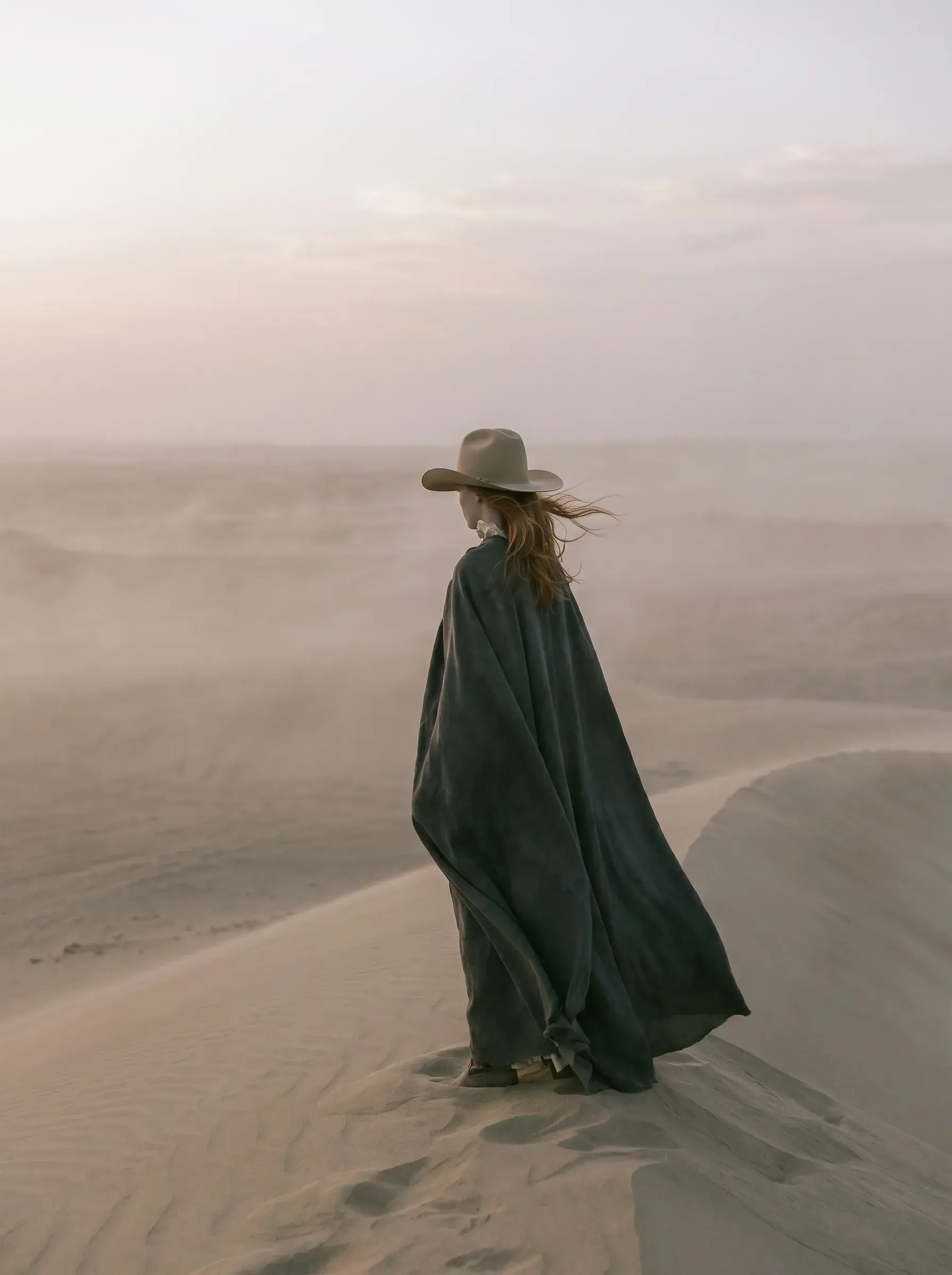 Femme de dos en grande cape sombre et chapeau debout sur une dune sous un ciel rose pâle
