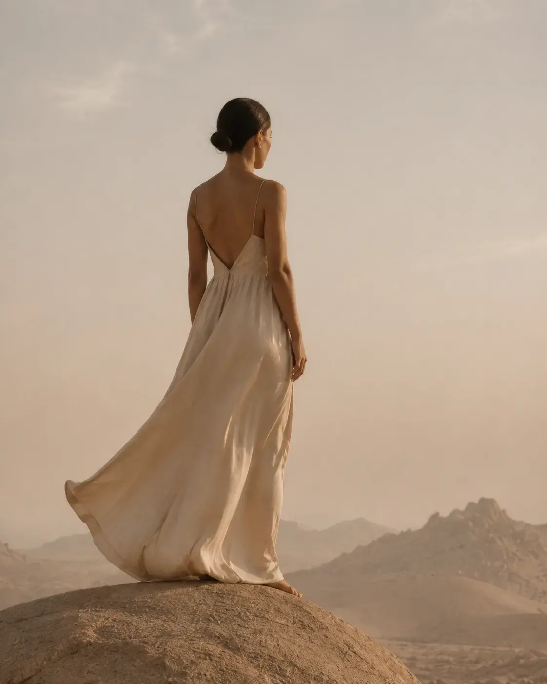 Femme de dos en robe blanche dos nu sur rocher – désert brumeux, montagnes, ciel pâle