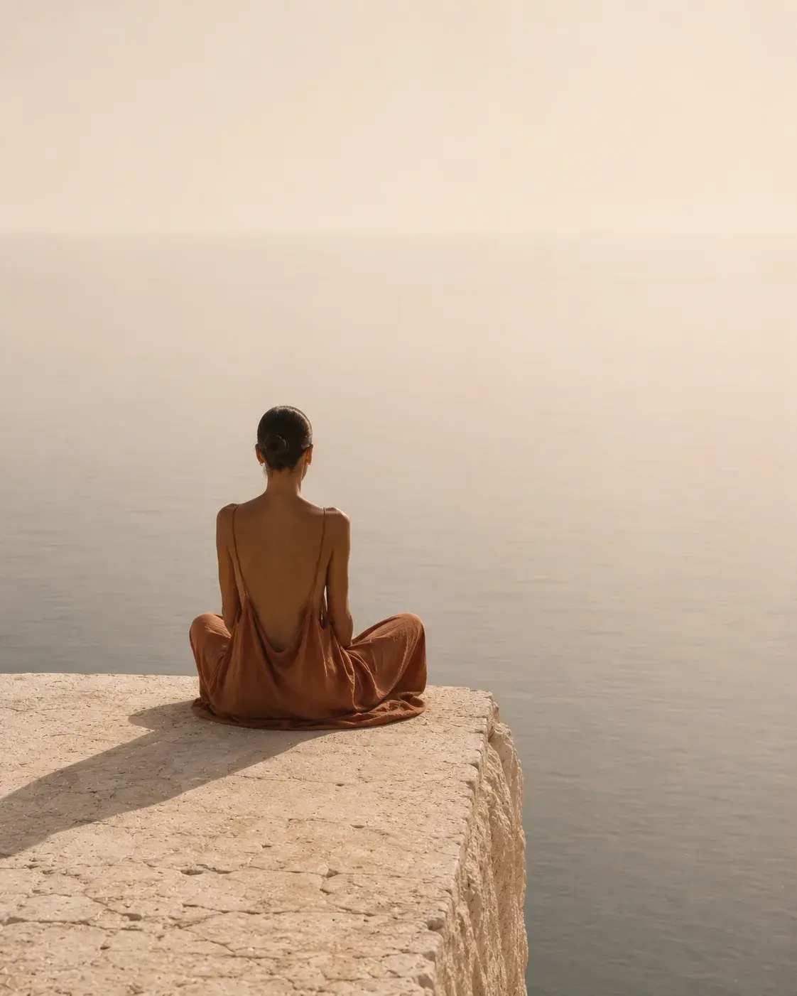 Femme de dos en robe satin terracotta en méditation sur falaise – mer brumeuse, horizon rosé
