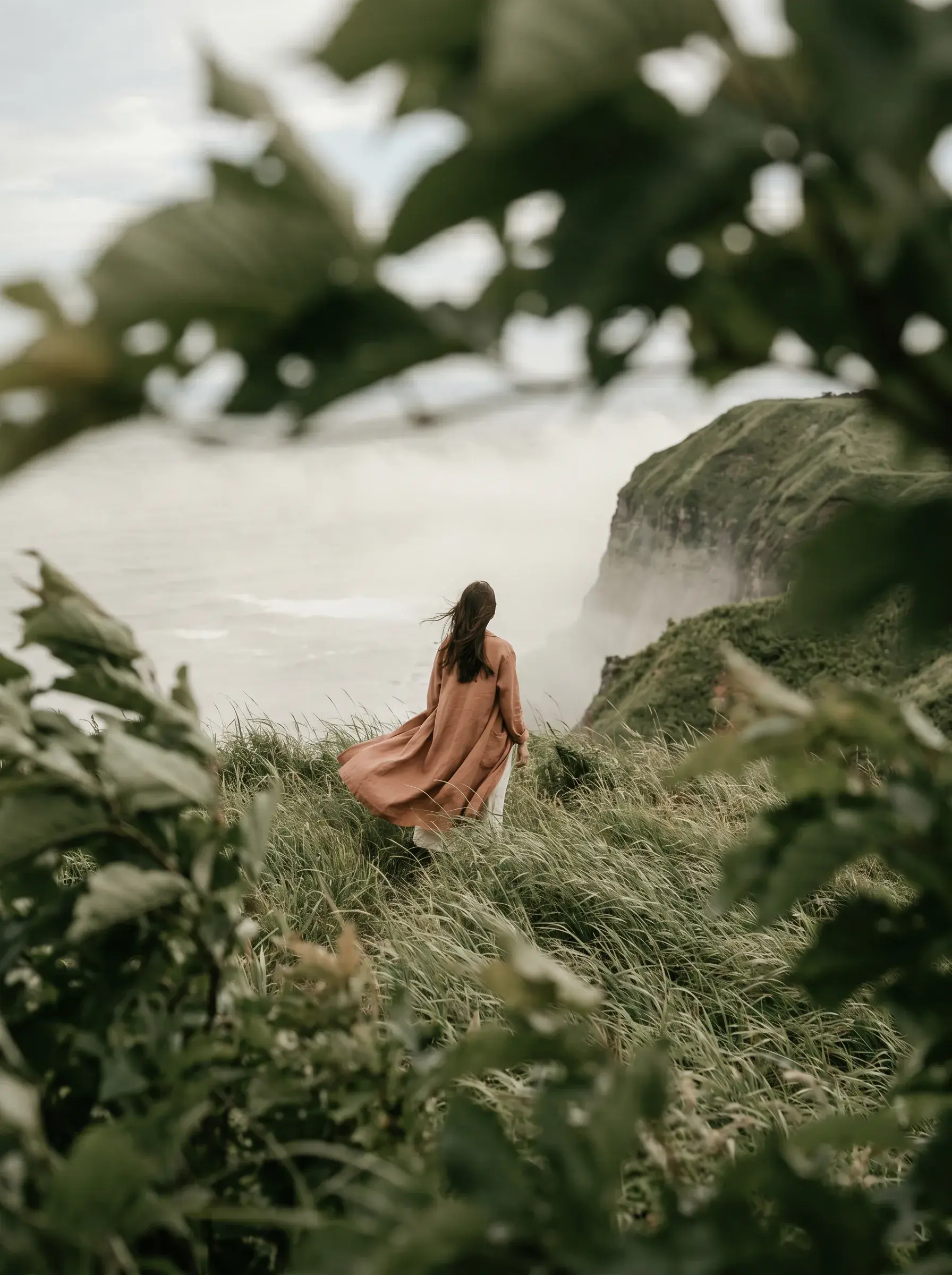 Femme de dos en robe terracotta dans herbes hautes – falaises côtières brumeuses en fond