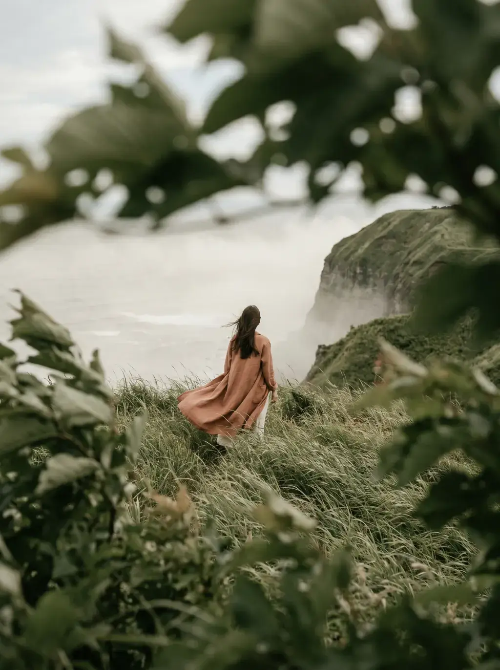 Femme de dos en robe terracotta dans herbes hautes – falaises côtières brumeuses en fond