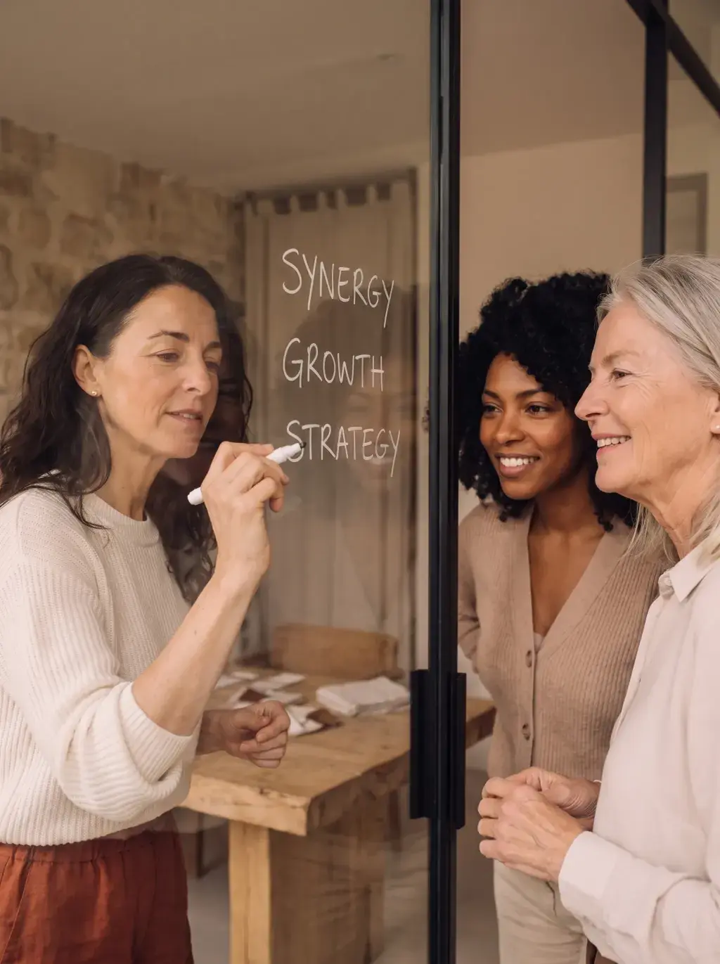 Femme écrivant Synergy Growth Strategy sur une vitre devant deux collègues souriantes