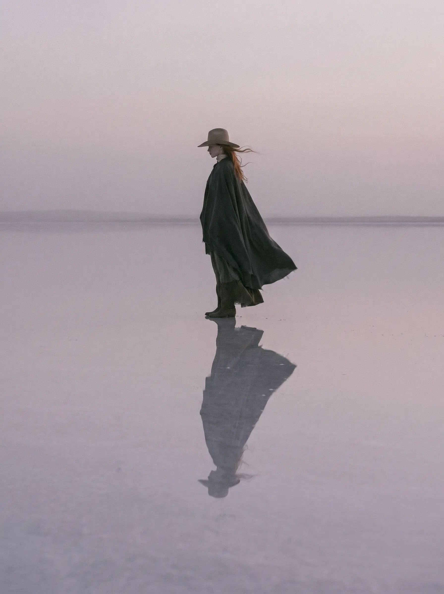 Femme en cape sombre et chapeau debout sur un lac salé rose avec reflet et vent dans un ciel rose