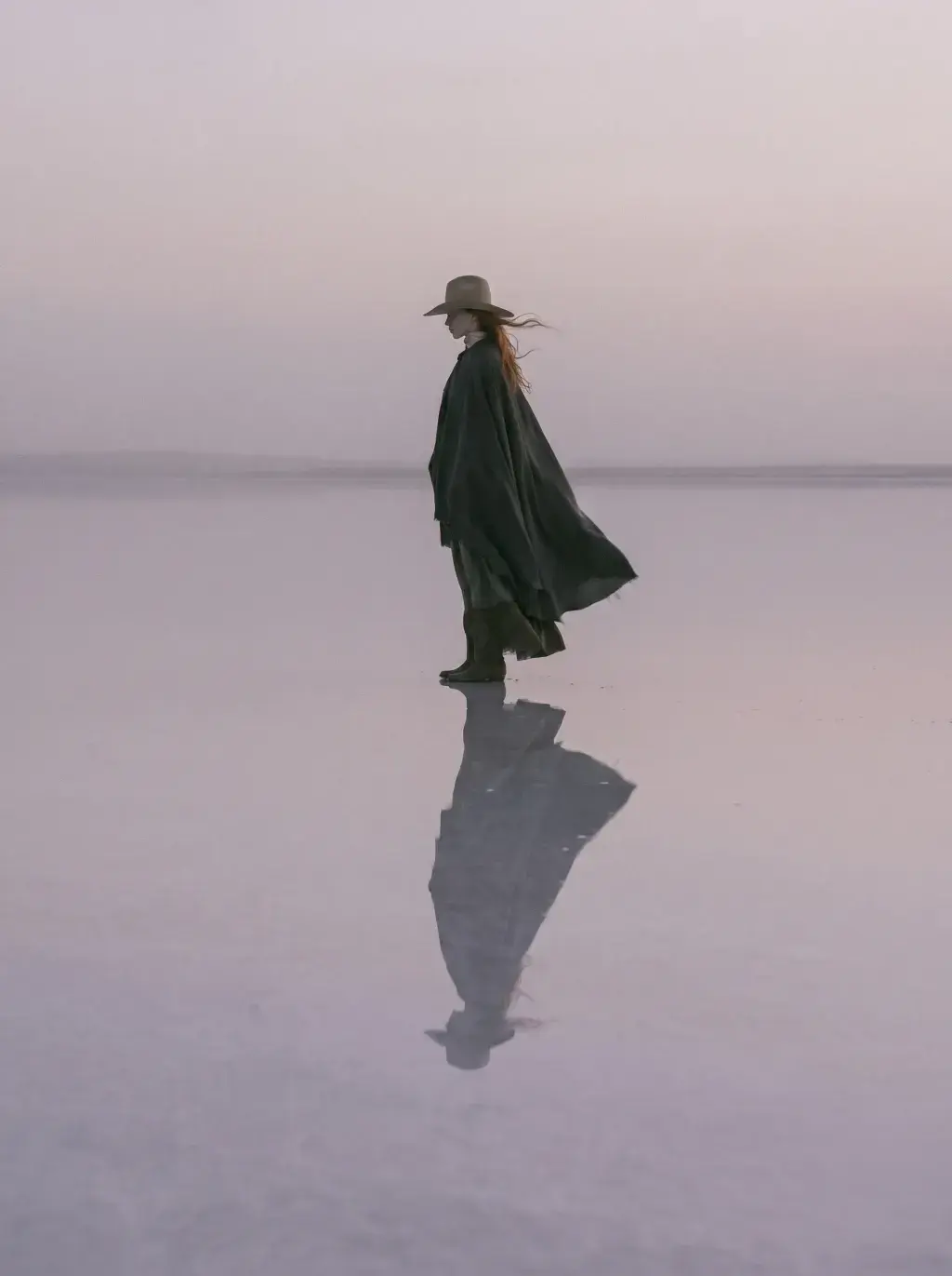 Femme en cape sombre et chapeau debout sur un lac salé rose avec reflet et vent dans un ciel rose