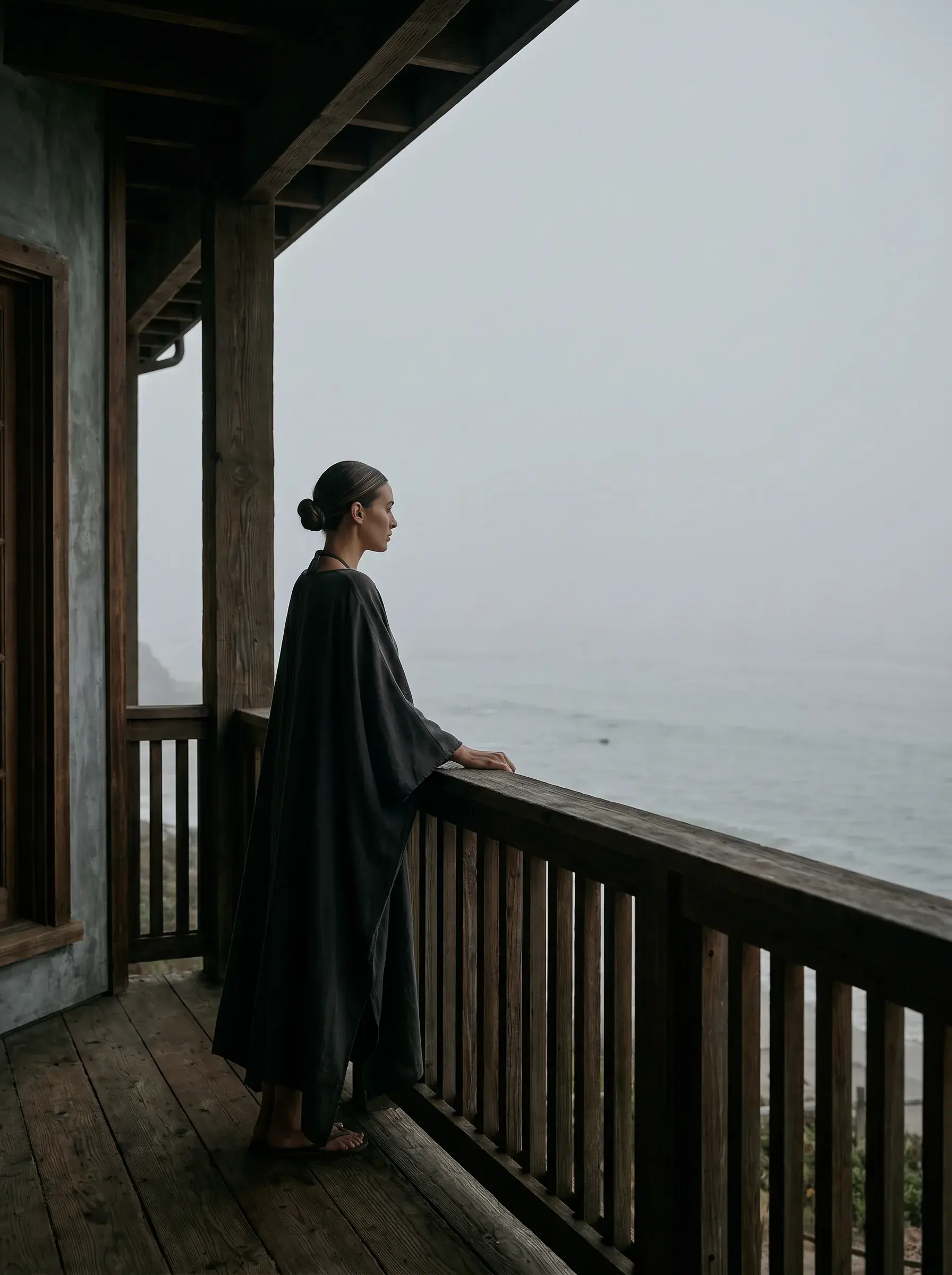 Femme en manteau sombre sur balcon en bois contemplant l'océan dans la brume