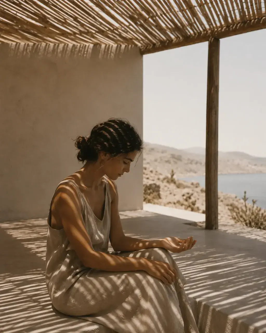 Femme en robe de lin assise sous pergola de roseau – jeu d'ombres rayées et vue mer