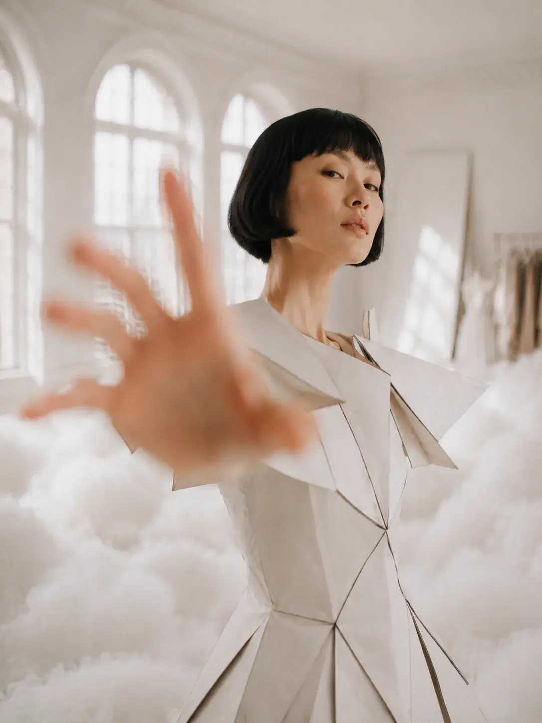 Femme en robe géométrique blanche tendant la main vers l'objectif – studio nuageux lumineux