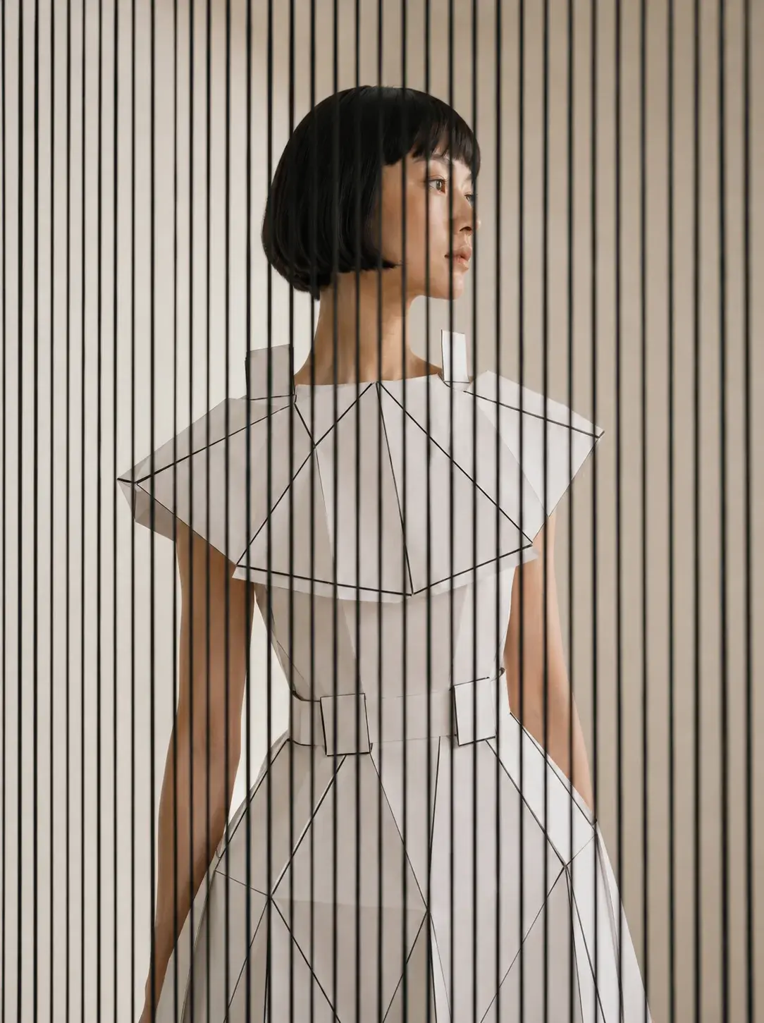 Femme en robe origami de profil devant panneau de lattes verticales – graphique et épuré
