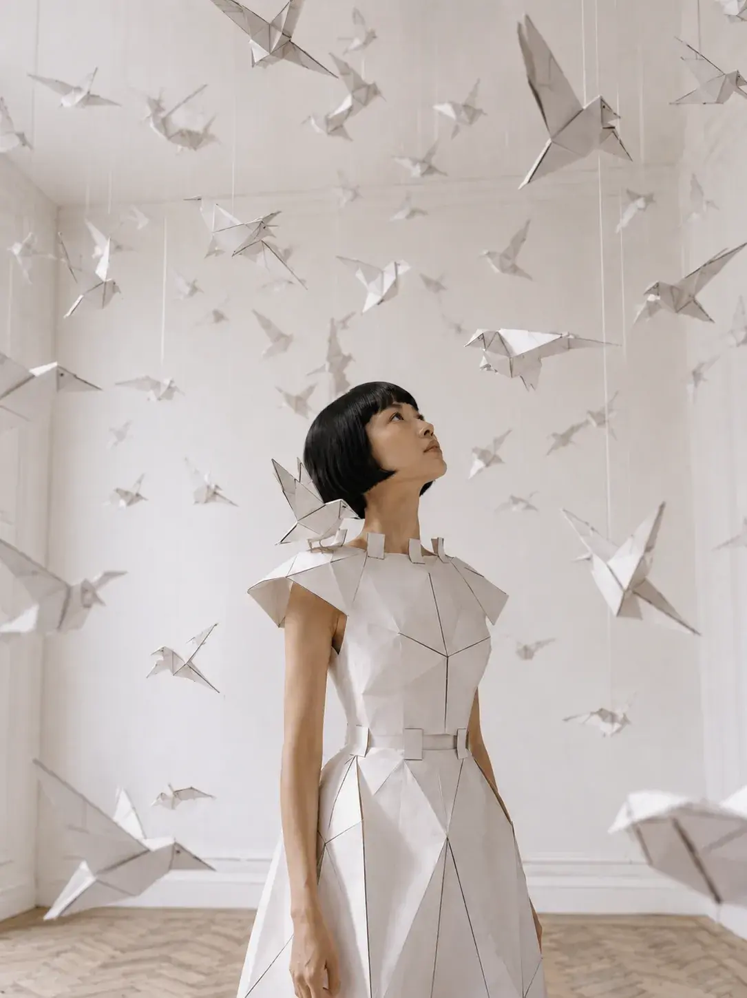 Femme en robe origami entourée de grues en papier suspendues – salon blanc et parquet
