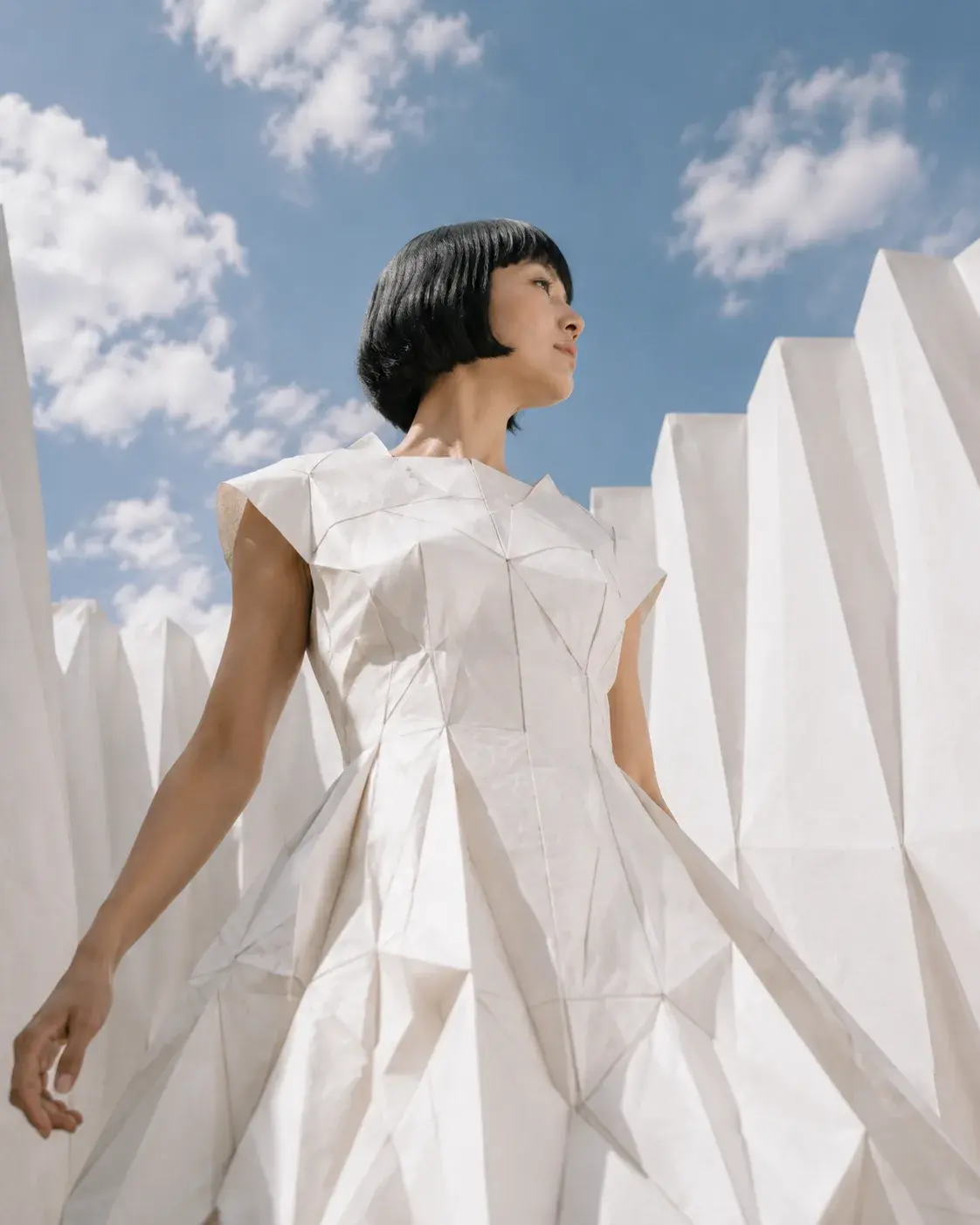 Femme en robe origami entre panneaux plissés blancs – contre-plongée ciel et nuages