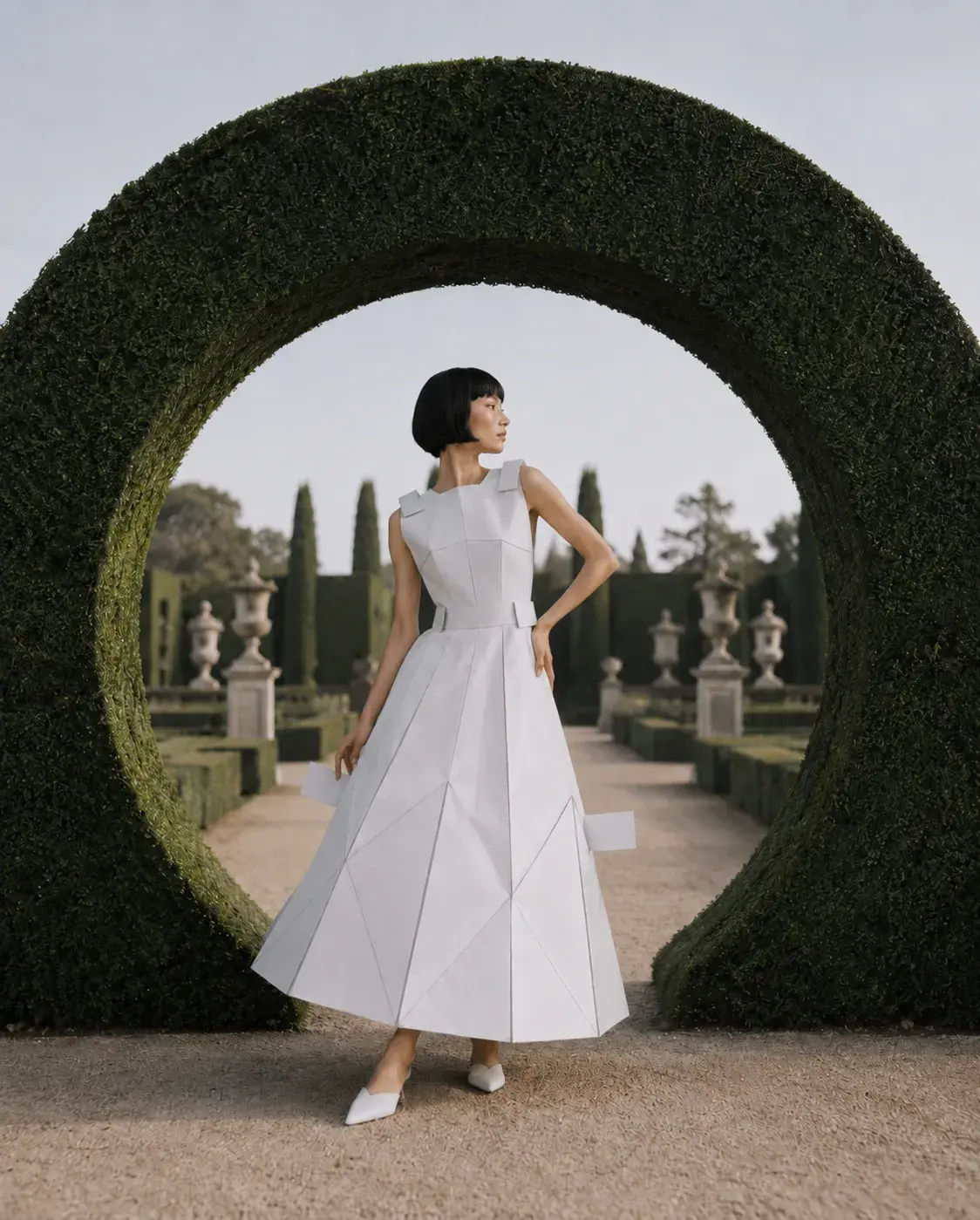 Femme en robe origami main sur hanche dans arche de buis – jardin à la française