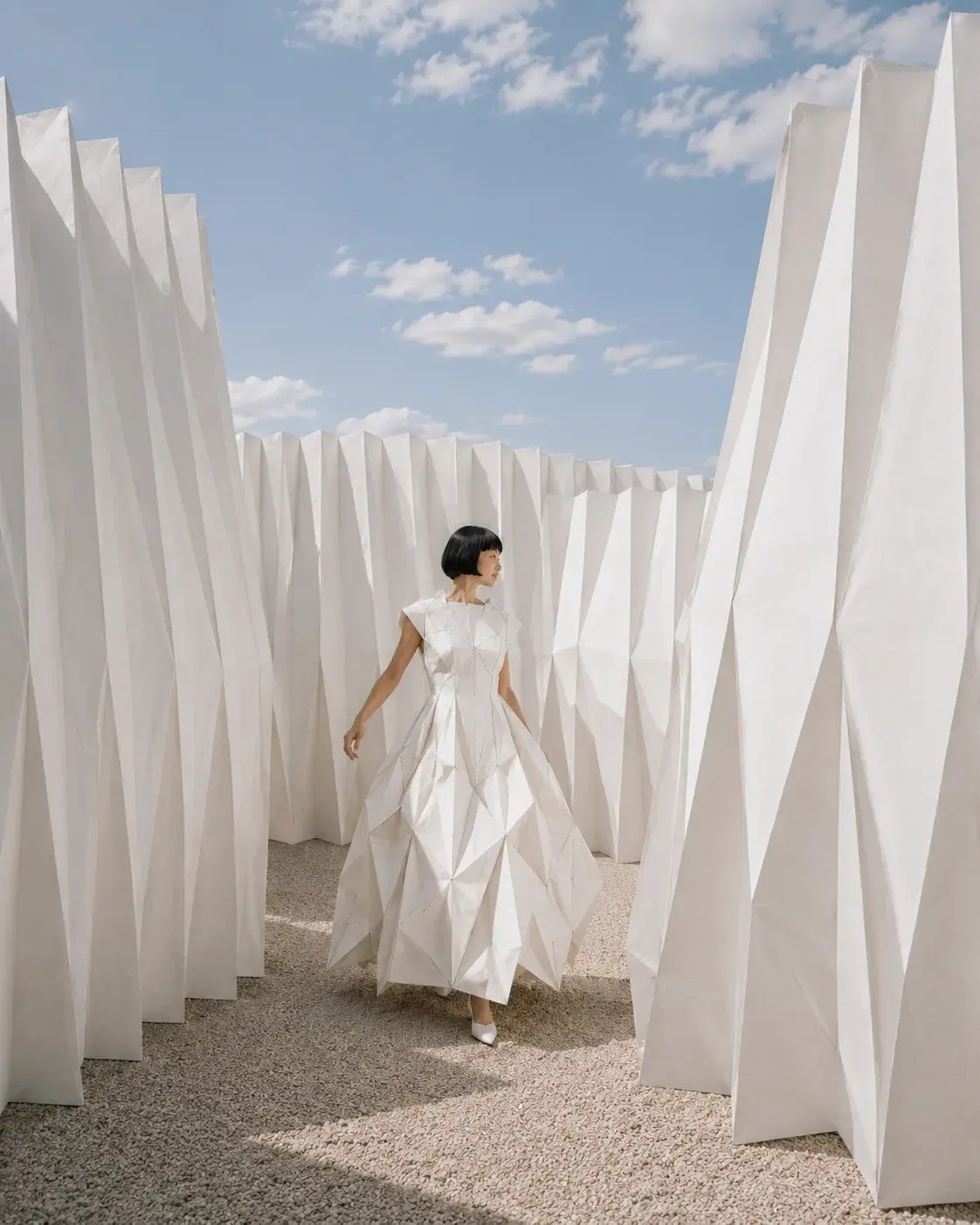 Femme en robe origami marchant dans labyrinthe de panneaux plissés blancs – ciel bleu