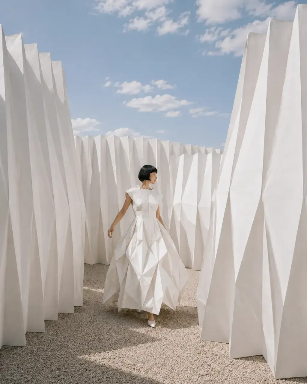 Femme en robe origami marchant dans labyrinthe de panneaux plissés blancs – ciel bleu