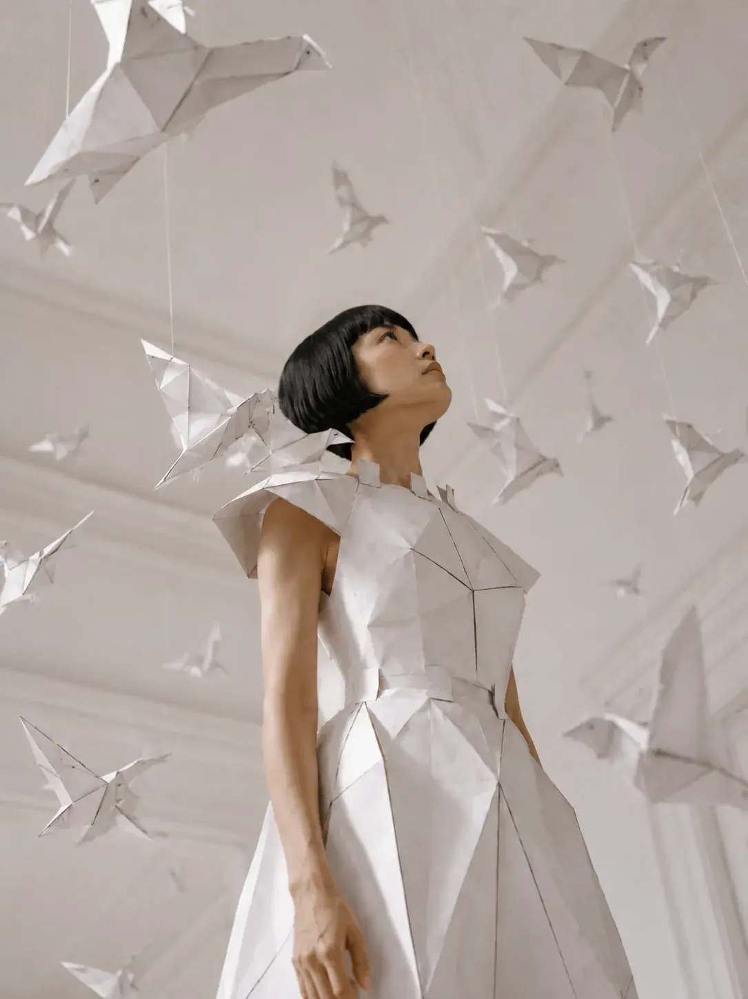 Femme en robe origami sous pluie de grues en papier – contre-plongée salon blanc