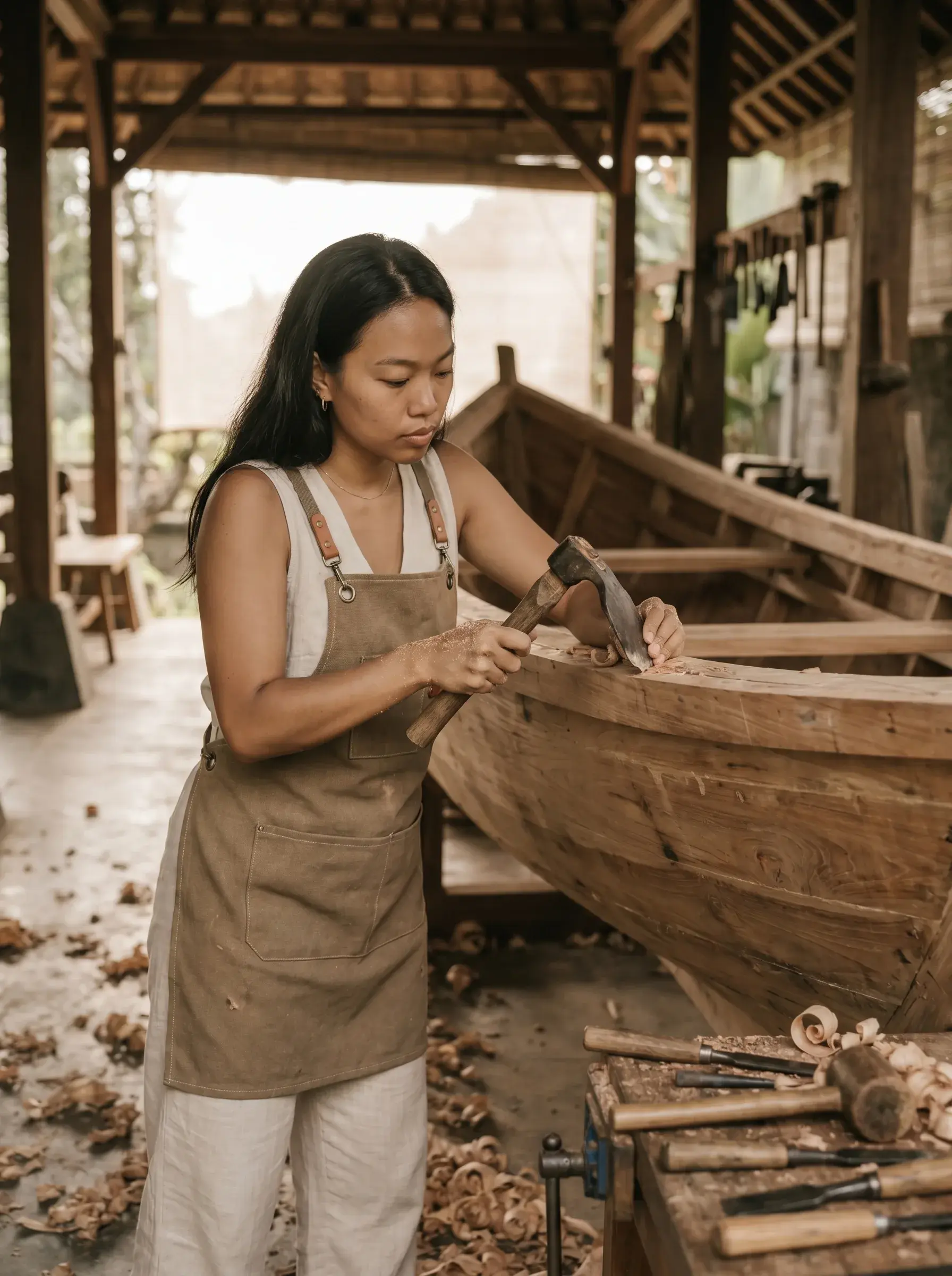 Femme en tablier lin travaillant la coque d'une barque en bois avec marteau et ciseau dans atelier balinais ouvert