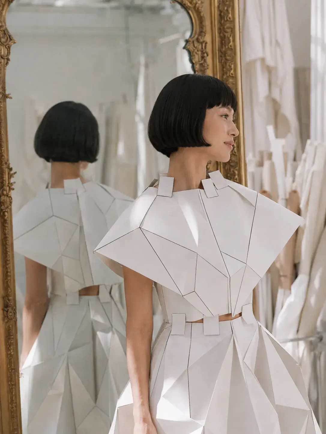 Femme en top origami devant miroir doré – reflet et portant de robes en fond