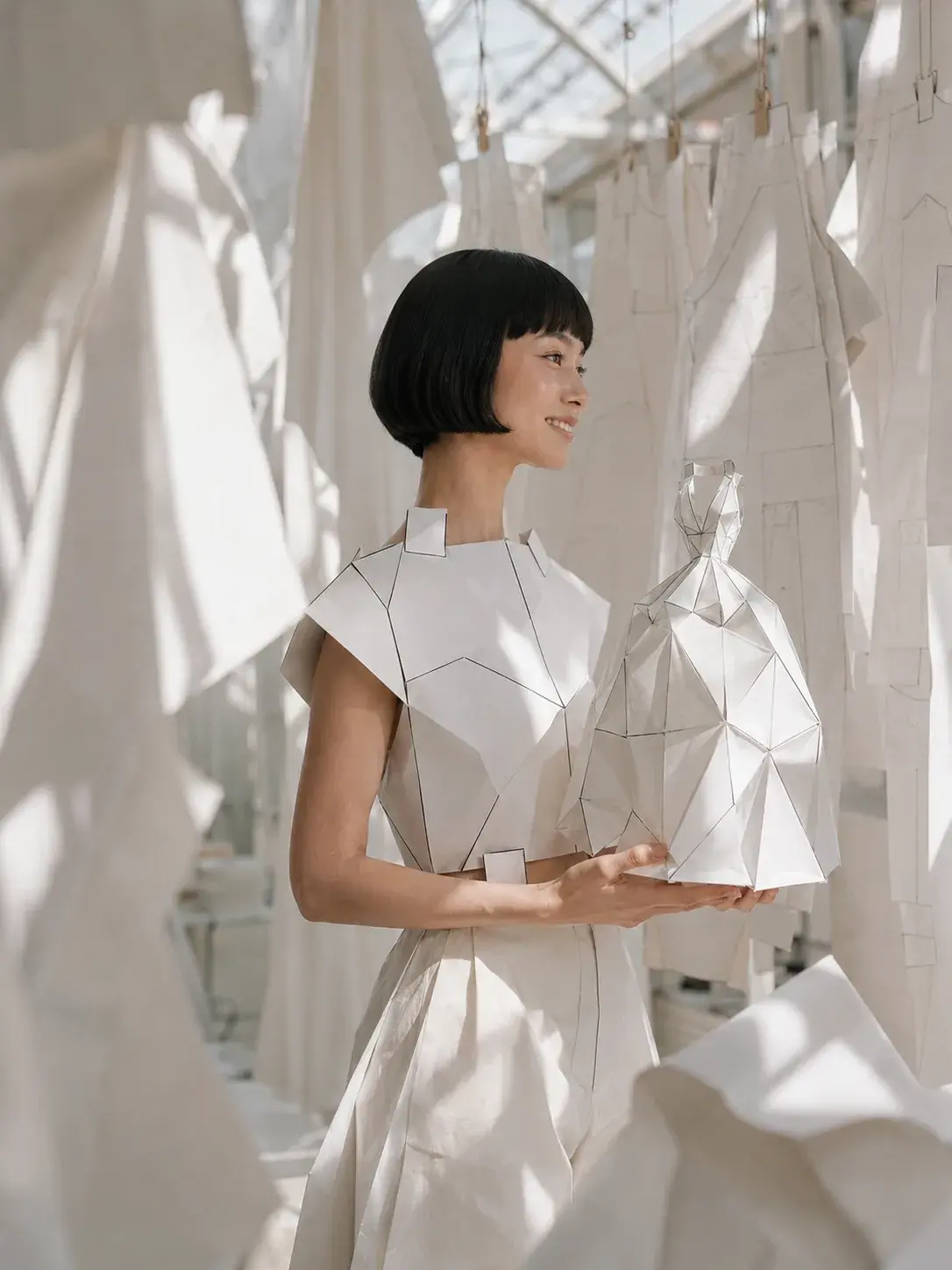 Femme en top origami souriante tenant un sac sculptural – textiles suspendus sous verrière
