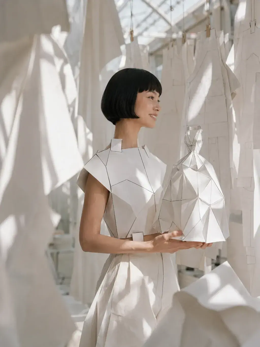 Femme en top origami souriante tenant un sac sculptural – textiles suspendus sous verrière