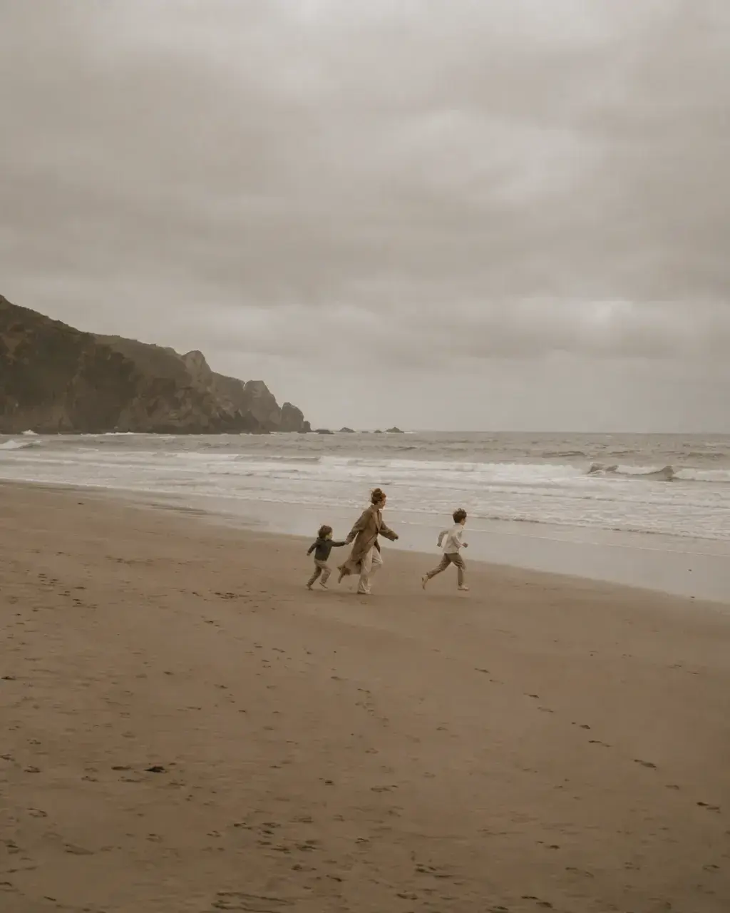 Femme et deux enfants courant sur une plage de sable – ciel nuageux, mer et falaises