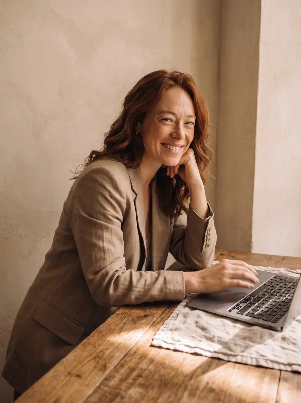 Femme rousse en blazer lin souriant devant son laptop sur une table rustique