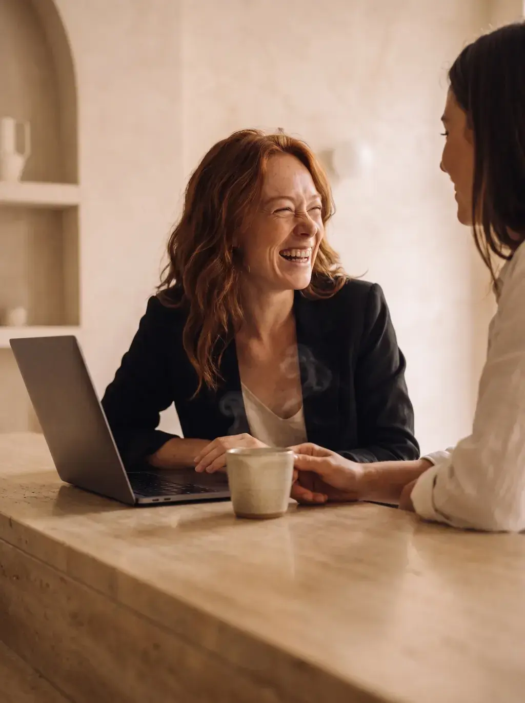 Femme rousse en blazer noir riant avec une collègue devant son laptop dans un café