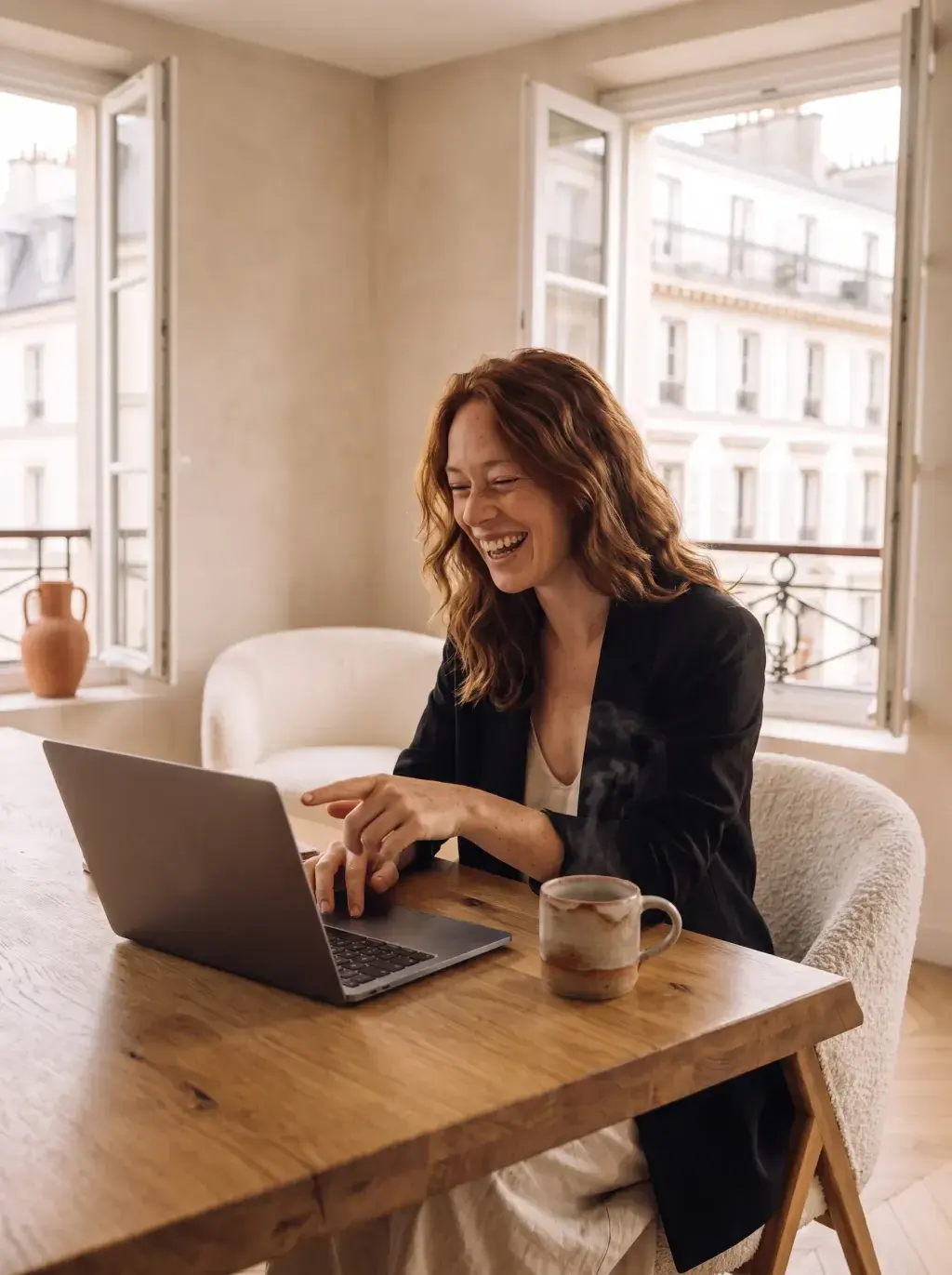 Femme rousse en blazer noir riant devant son laptop dans un appartement parisien
