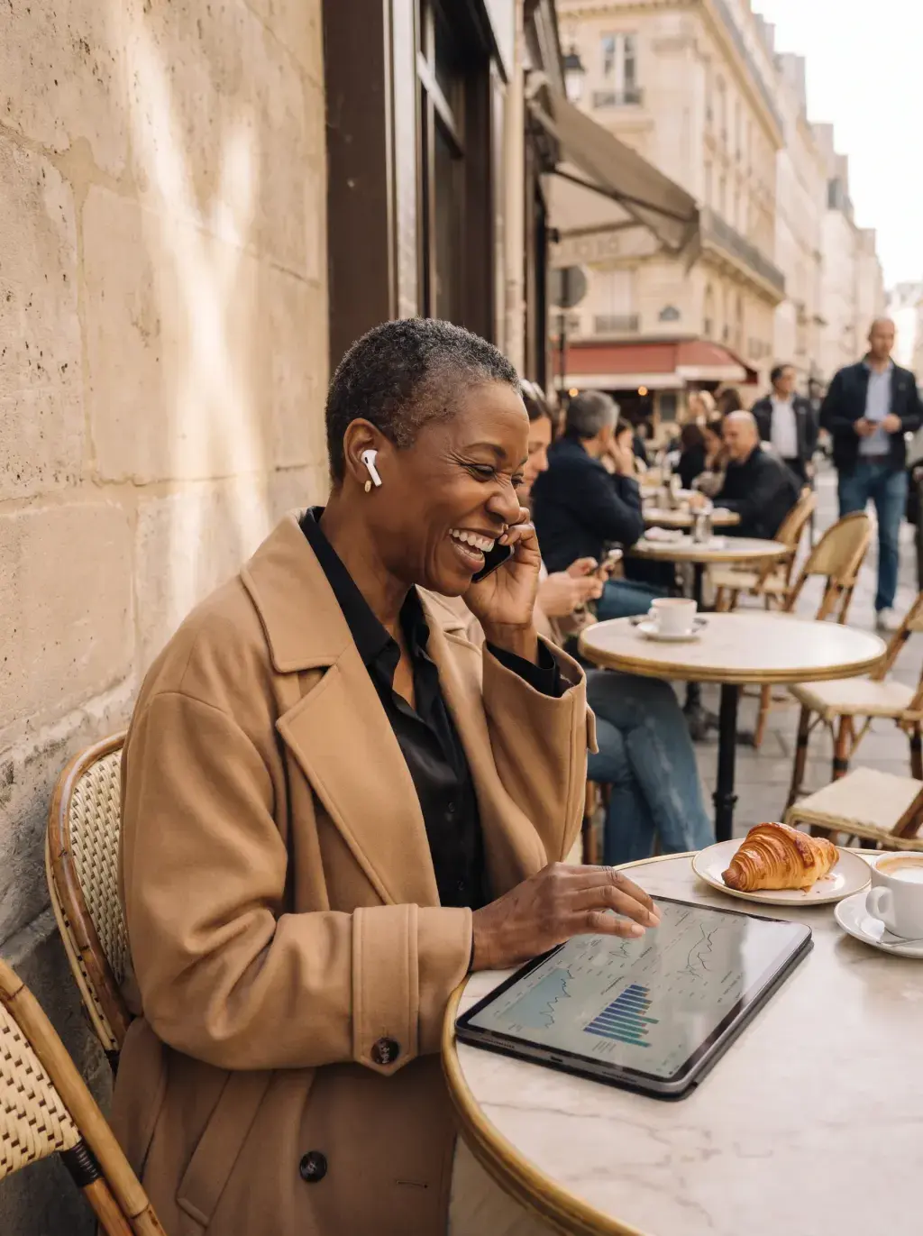 Femme senior noire riant au téléphone en terrasse animée à Paris avec tablette