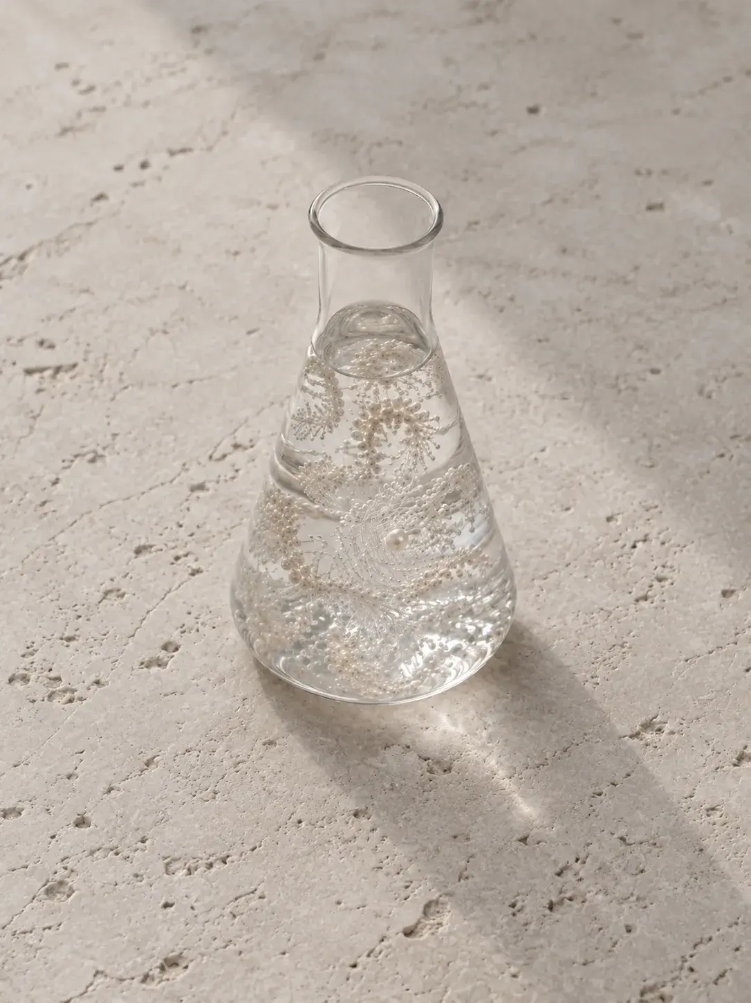 Fiole Erlenmeyer en verre sur travertin, collier de perles en suspension dans un liquide clair