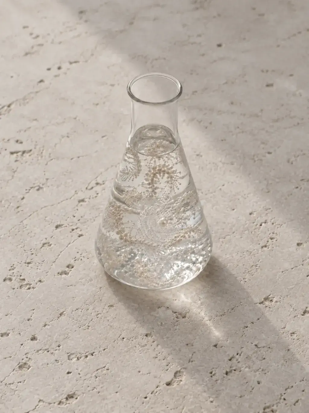 Fiole Erlenmeyer en verre sur travertin, collier de perles en suspension dans un liquide clair