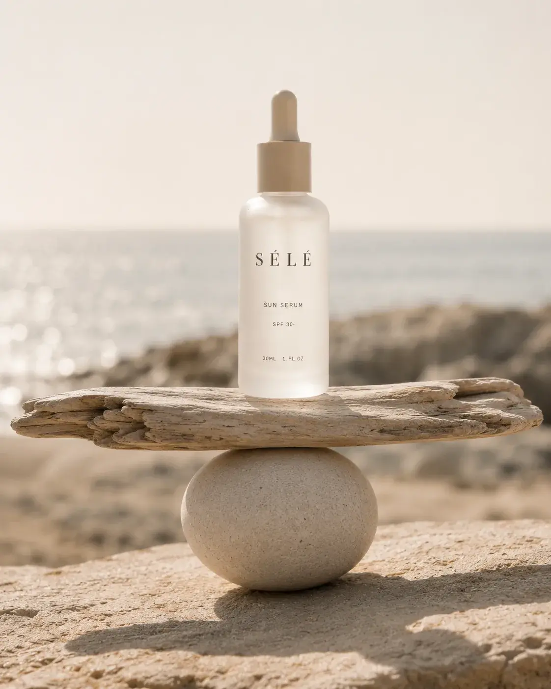 Flacon sérum en équilibre sur bois flotté et galet – plage, mer en fond, composition zen