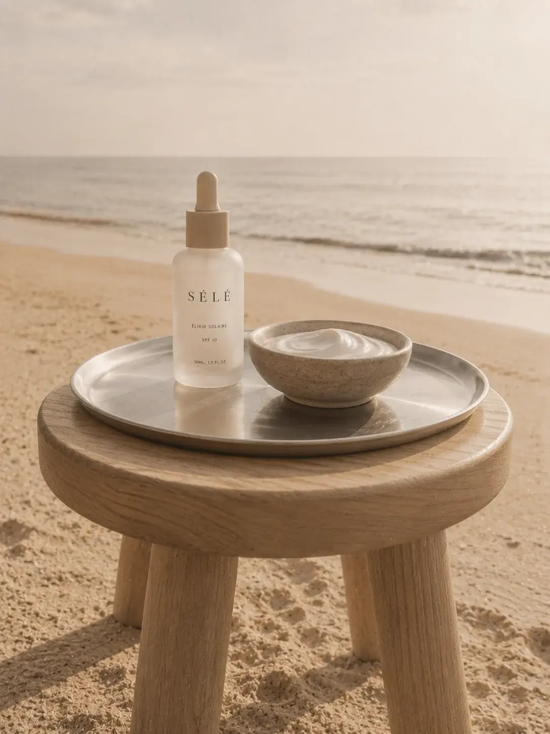 Flacon sérum et bol crème sur plateau métal – tabouret bois sur plage, horizon mer, coucher de soleil