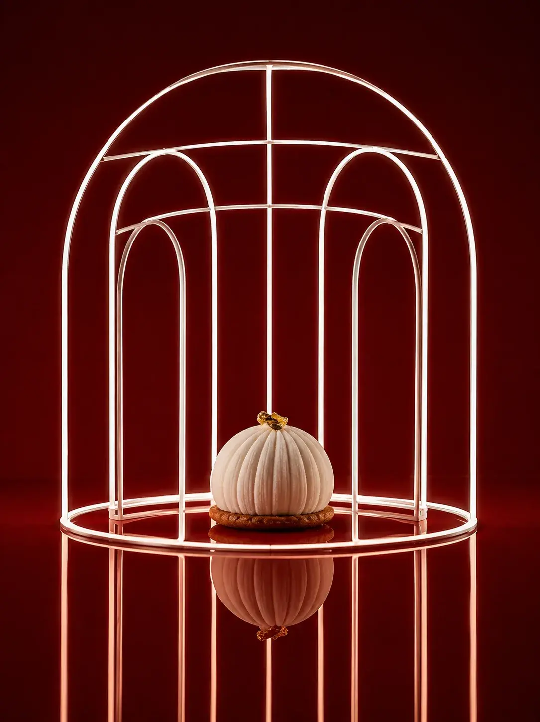 Gâteau citrouille côtelée blanche sur socle doré sous cage architecturale en fils dorés, table-miroir rouge avec reflet