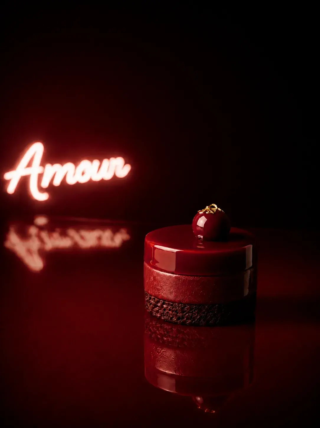 Gâteau individuel rouge cylindrique avec topping cerise dorée devant néon « Amour » flou, reflet sur surface miroir