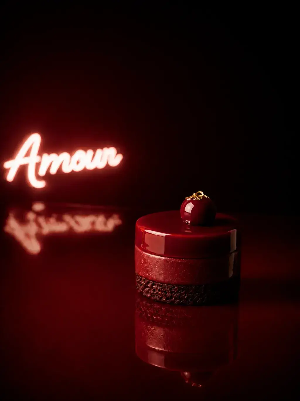 Gâteau individuel rouge cylindrique avec topping cerise dorée devant néon « Amour » flou, reflet sur surface miroir