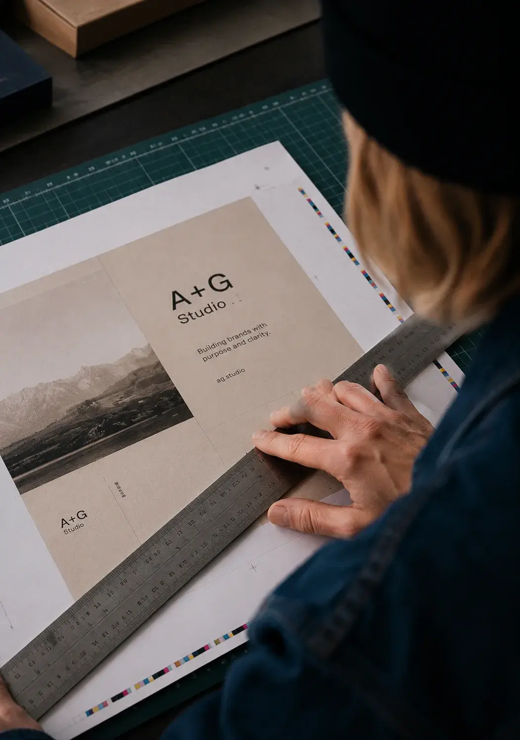 Graphiste de dos guidant une règle métallique sur la mise en page A+G Studio étalée sur tapis de découpe vert