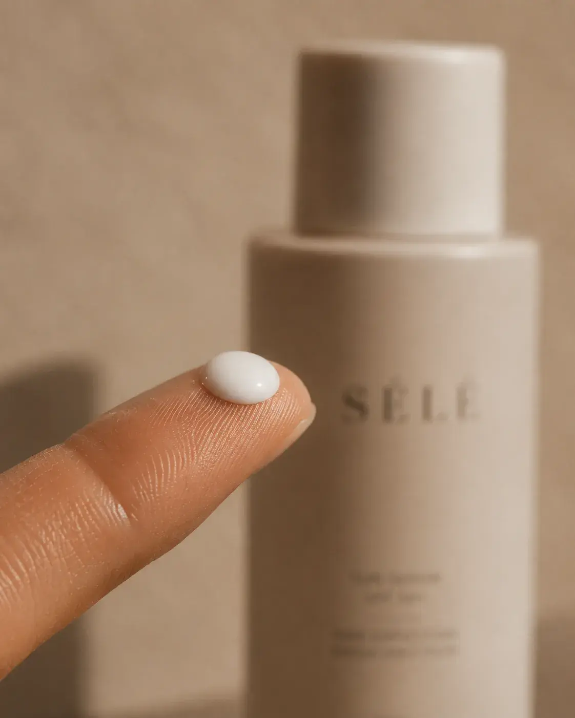 Gros plan doigt index avec perle de crème blanche – flacon beige flou, fond neutre
