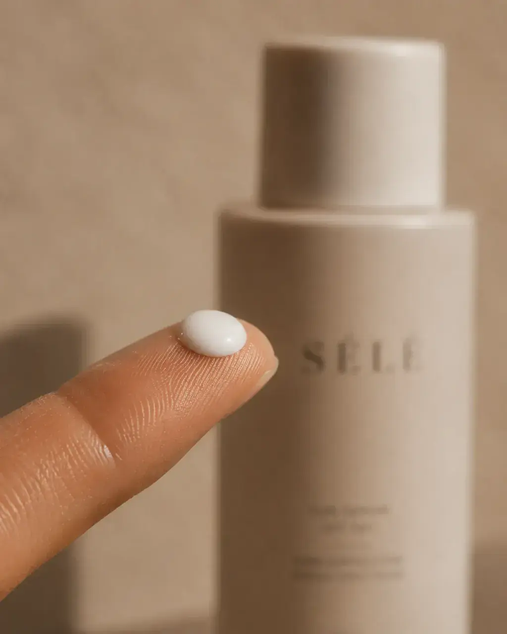 Gros plan doigt index avec perle de crème blanche – flacon beige flou, fond neutre